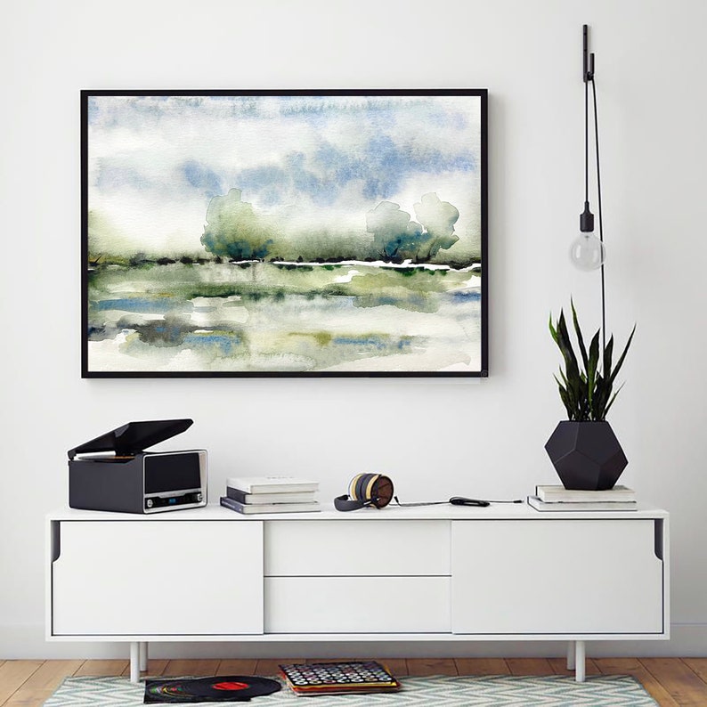 Op de afbeelding: Een ingelijst aquarel schilderij van een landschap met een meer en bomen. Het schilderij hangt aan een witte muur boven een witte kast met een platenspeler, boeken en koptelefoon erop.