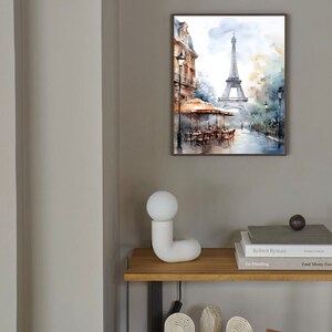 K&ouml;nnte beinhalten: Ein gerahmtes Aquarellbild des Eiffelturms in Paris, Frankreich. Das Bild zeigt eine Stra&szlig;enszene mit einem Caf&eacute; und Passanten. Das Bild h&auml;ngt an einer hellgrauen Wand &uuml;ber einem Holzregal mit einer wei&szlig;en Keramiklampe und B&uuml;chern.