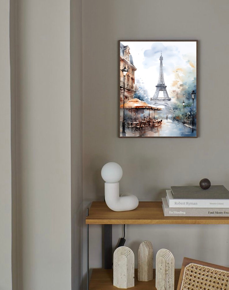 K&ouml;nnte beinhalten: Ein gerahmtes Aquarellbild des Eiffelturms in Paris, Frankreich. Das Bild zeigt eine Stra&szlig;enszene mit einem Caf&eacute; und Passanten. Das Bild h&auml;ngt an einer hellgrauen Wand &uuml;ber einem Holzregal mit einer wei&szlig;en Keramiklampe und B&uuml;chern.