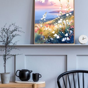 Pode incluir: Uma pintura em aquarela emoldurada retrata uma paisagem costeira com flores silvestres brancas em primeiro plano. A obra de arte apresenta um p&ocirc;r do sol com tons rosa e azul e uma costa distante. A pintura &eacute; exibida em uma parede cinza.