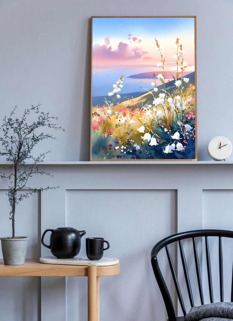 Pode incluir: Uma pintura em aquarela emoldurada retrata uma paisagem costeira com flores silvestres brancas em primeiro plano. A obra de arte apresenta um p&ocirc;r do sol com tons rosa e azul e uma costa distante. A pintura &eacute; exibida em uma parede cinza.