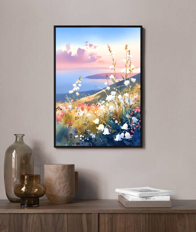 Pode incluir: Uma impress&atilde;o em aquarela emoldurada de uma paisagem. A obra de arte retrata uma colina com flores brancas em forma de sino, flores silvestres coloridas e uma vista do mar sob um c&eacute;u rosa e azul. A impress&atilde;o &eacute; exibida em uma superf&iacute;cie de madeira.