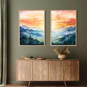 May include: Two framed watercolor landscape prints. Each features a mountain range with blue and green hues, a forest, and a sunset with orange and yellow tones. The prints are displayed above a wooden cabinet.