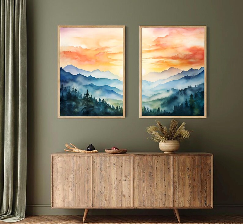 May include: Two framed watercolor landscape prints. Each features a mountain range with blue and green hues, a forest, and a sunset with orange and yellow tones. The prints are displayed above a wooden cabinet.