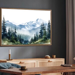 May include: A rectangular wall art piece featuring a watercolour landscape painting of a mountain range with snow-capped peaks and a dense forest of dark green and blue pine trees in the foreground, all enclosed within a thin light wood frame.