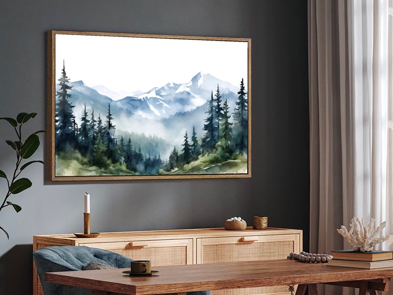 May include: A rectangular wall art piece featuring a watercolour landscape painting of a mountain range with snow-capped peaks and a dense forest of dark green and blue pine trees in the foreground, all enclosed within a thin light wood frame.
