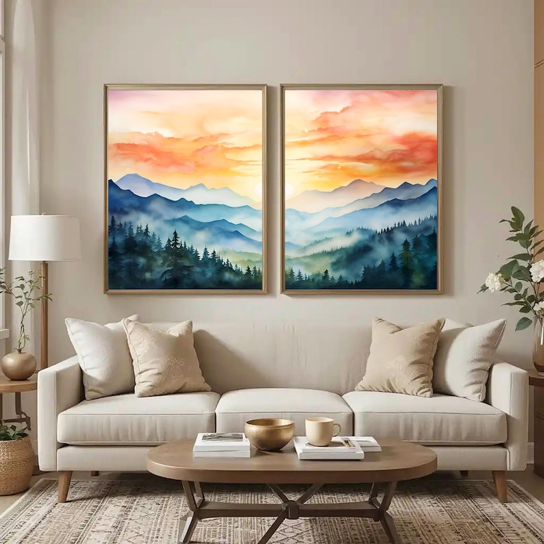 May include: Two framed watercolor landscape paintings. The artwork depicts layered blue mountains and green forests under a vibrant orange and yellow sunset sky. The paintings are displayed above a beige sofa in a living room setting.