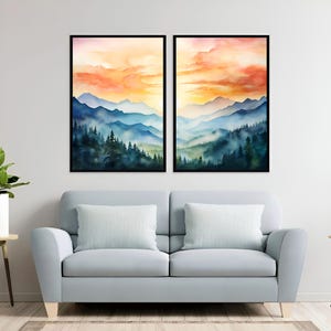 May include: Two framed watercolor landscape paintings. Each features a mountain range scene with layers of blue and green mountains, a forest, and a sunset with orange and yellow hues. The paintings are displayed above a light blue sofa with white pillows.