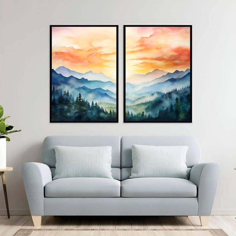 May include: Two framed watercolor landscape paintings. Each features a mountain range scene with layers of blue and green mountains, a forest, and a sunset with orange and yellow hues. The paintings are displayed above a light blue sofa with white pillows.