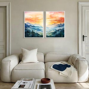 May include: Two framed watercolor paintings of mountain landscapes. The artwork features layers of blue mountains under an orange and yellow sunset. The paintings are displayed above a cream-colored sofa in a modern living room.