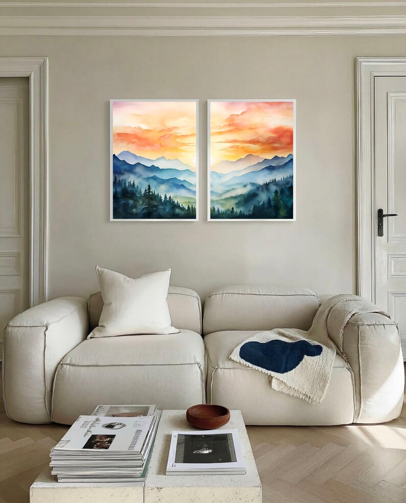 May include: Two framed watercolor paintings of mountain landscapes. The artwork features layers of blue mountains under an orange and yellow sunset. The paintings are displayed above a cream-colored sofa in a modern living room.