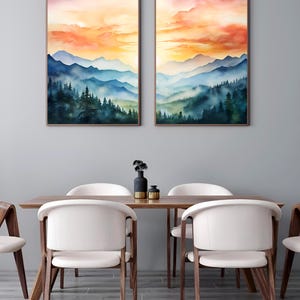 May include: Two framed watercolor landscape paintings depict a mountain range under a vibrant sunset sky. The artwork features layers of blue mountains and green trees. The paintings are displayed above a wooden dining table with white chairs.