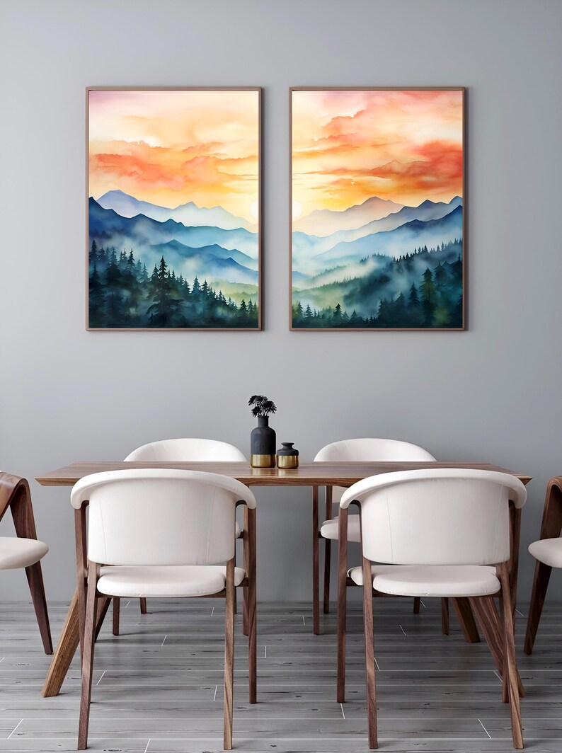 May include: Two framed watercolor landscape paintings depict a mountain range under a vibrant sunset sky. The artwork features layers of blue mountains and green trees. The paintings are displayed above a wooden dining table with white chairs.