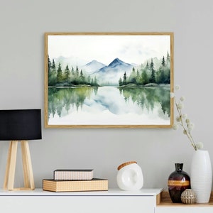 May include: A watercolor painting of a lake with mountains and trees reflected in the water. The painting is framed in a light wood frame.