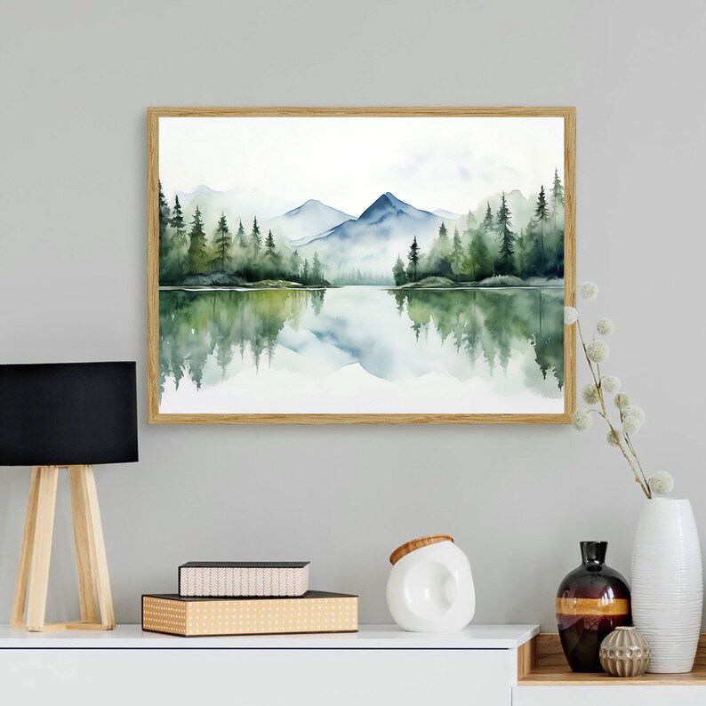 May include: A watercolor painting of a lake with mountains and trees reflected in the water. The painting is framed in a light wood frame.