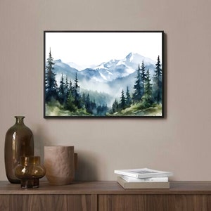 May include: A rectangular wall art print in a black frame featuring a watercolour landscape painting of snow-capped mountains rising above a dense forest of dark green and blue pine trees with soft mist settling between the trees and peaks.