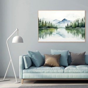 May include: A watercolor painting of a mountain lake scene with a reflection of the trees and mountains in the water. The painting is framed in a light wood frame and is hanging on a light gray wall above a light blue sofa with brown and gray throw pillows.
