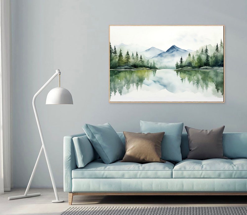 May include: A watercolor painting of a mountain lake scene with a reflection of the trees and mountains in the water. The painting is framed in a light wood frame and is hanging on a light gray wall above a light blue sofa with brown and gray throw pillows.