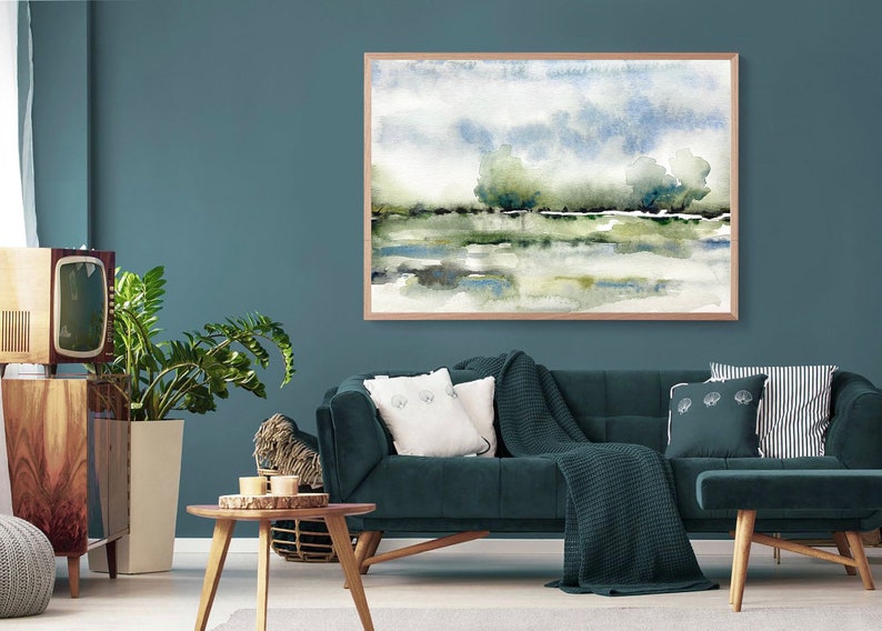 Op de afbeelding: Een tealblauwe woonkamer met een donkerblauwe fluwelen bank, een houten salontafel, een plant in een pot en een ingelijst aquarel schilderij van een landschap met bomen en water.