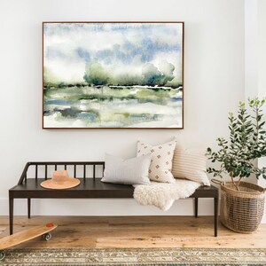 Op de afbeelding: Een aquarel schilderij van een landschap met bomen en een waterpartij. Het schilderij is ingelijst in een lichtbruin lijst en hangt aan een witte muur boven een zwarte bank.