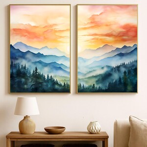 May include: Two framed watercolor landscape paintings. Each features a mountain range with a forest in the foreground. The sky is a gradient of orange, yellow, and red. The paintings are in gold frames.