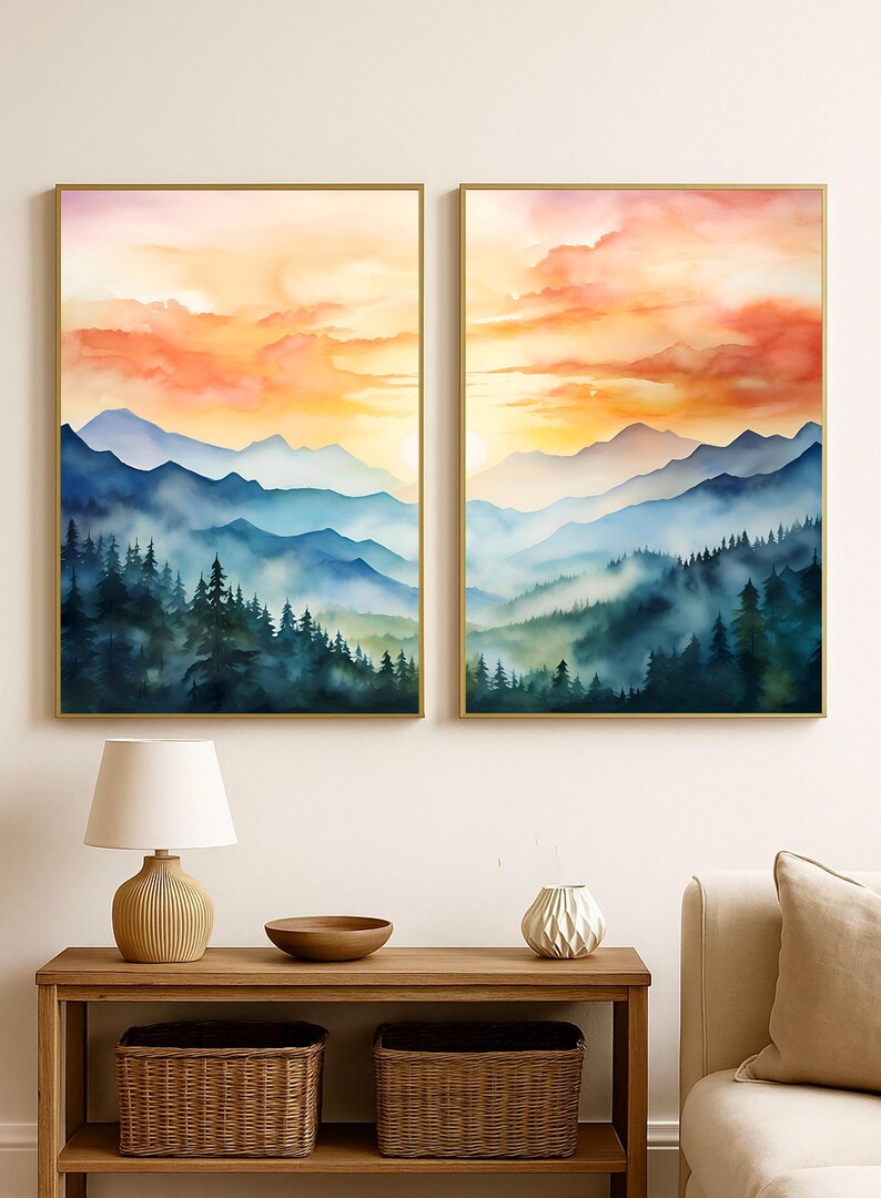 May include: Two framed watercolor landscape paintings. Each features a mountain range with a forest in the foreground. The sky is a gradient of orange, yellow, and red. The paintings are in gold frames.
