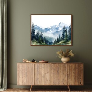 May include: A rectangular wall art print featuring a watercolour landscape of snow-capped mountains and a dense forest of dark green and blue pine trees. The scene depicts misty valleys and layered peaks in shades of blue, white, and muted green.