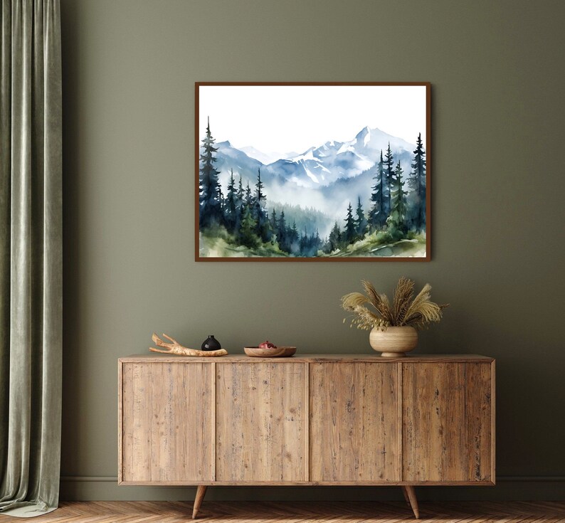 May include: A rectangular wall art print featuring a watercolour landscape of snow-capped mountains and a dense forest of dark green and blue pine trees. The scene depicts misty valleys and layered peaks in shades of blue, white, and muted green.