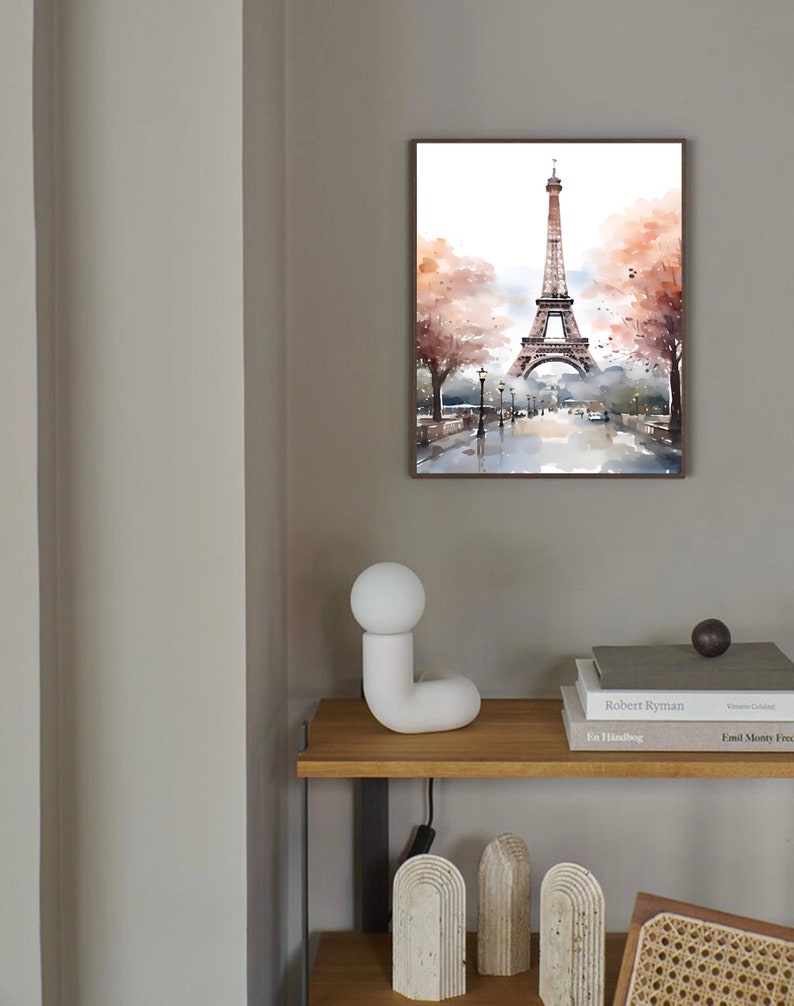 May include: A watercolor painting of the Eiffel Tower in Paris, France. The painting is framed and hanging on a light gray wall. There is a white ceramic lamp on a wooden shelf below the painting.