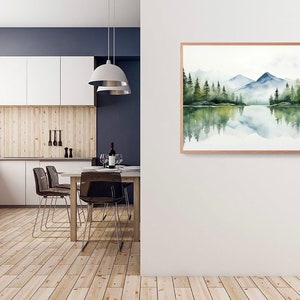 May include: A watercolor painting of a mountain lake scene with a reflection of the mountains and trees in the water. The painting is framed in a light wood frame and is hanging on a white wall.