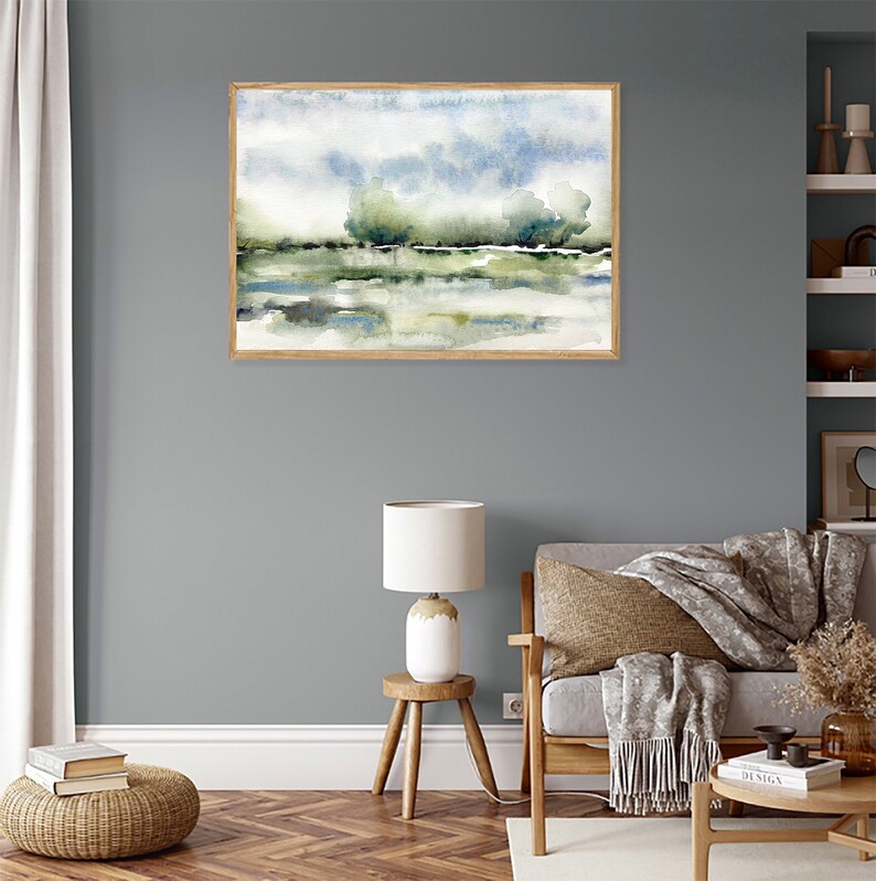 Op de afbeelding: Een ingelijst aquarel schilderij van een landschap met een blauwe lucht, groene bomen en een waterpartij. Het schilderij hangt aan een grijze muur in een woonkamer.