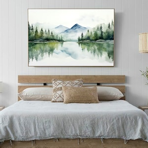 May include: A watercolor painting of a lake with mountains and trees in the background. The lake is reflecting the mountains and trees. The painting is framed and hanging on a white wall above a bed.