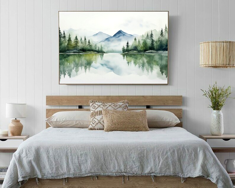 May include: A watercolor painting of a lake with mountains and trees in the background. The lake is reflecting the mountains and trees. The painting is framed and hanging on a white wall above a bed.