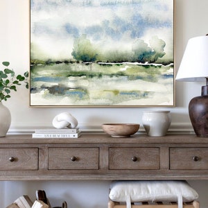 Op de afbeelding: Een aquarel schilderij van een landschap met bomen en een waterpartij. Het schilderij is ingelijst in een gouden lijst en hangt aan een witte muur boven een houten consoletafel.