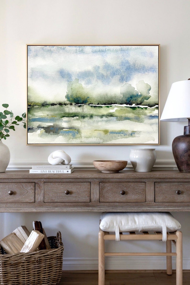Op de afbeelding: Een aquarel schilderij van een landschap met bomen en een waterpartij. Het schilderij is ingelijst in een gouden lijst en hangt aan een witte muur boven een houten consoletafel.