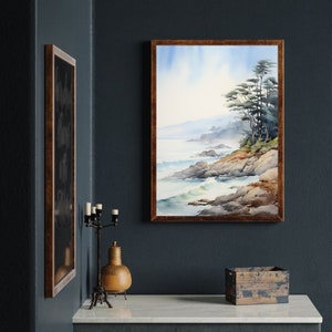 May include: Watercolor painting of a coastal landscape with a rocky shoreline, ocean waves, and trees. The artwork is framed in a dark wood frame. The painting is displayed on a dark blue wall, with a decorative candle holder and a wooden box on a white surface.