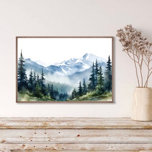May include: A framed watercolour landscape painting depicting a mountain range with snow-capped peaks, dense evergreen forests, and misty valleys in shades of blue, green, and white. The artwork is displayed in a thin brown wooden frame.
