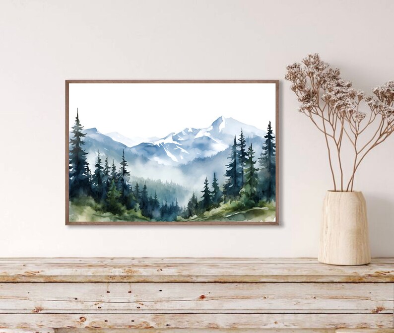 May include: A framed watercolour landscape painting depicting a mountain range with snow-capped peaks, dense evergreen forests, and misty valleys in shades of blue, green, and white. The artwork is displayed in a thin brown wooden frame.