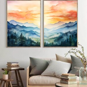 May include: Two framed watercolor paintings depict a mountain landscape with a sunset. The artwork features layers of blue mountains, green trees, and an orange and yellow sky. The paintings are displayed above a sofa with pillows.