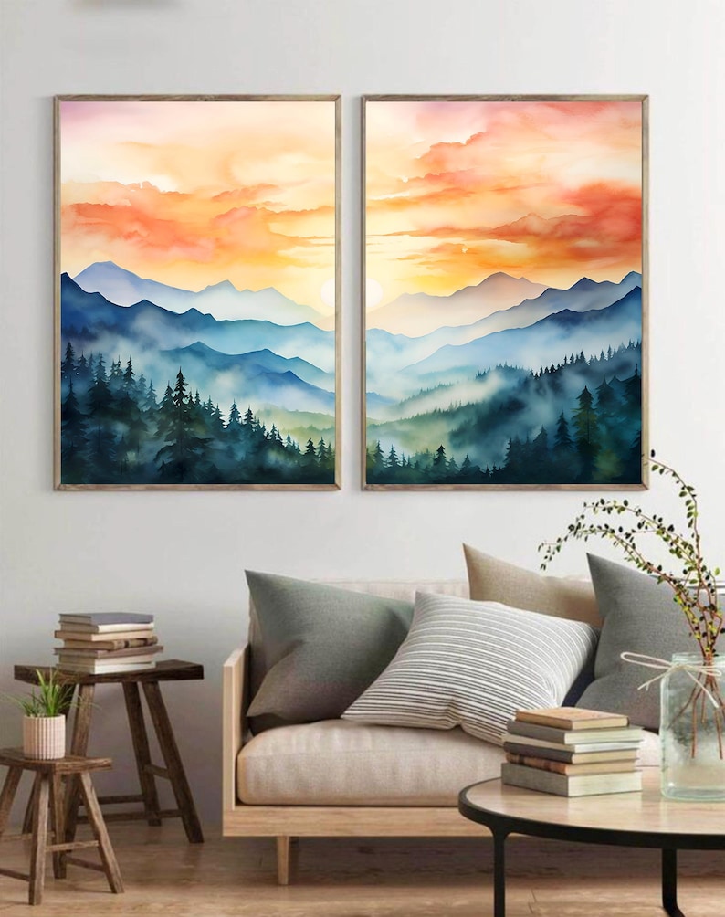 May include: Two framed watercolor paintings depict a mountain landscape with a sunset. The artwork features layers of blue mountains, green trees, and an orange and yellow sky. The paintings are displayed above a sofa with pillows.