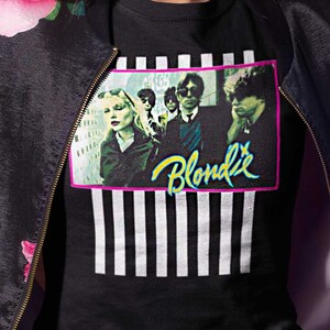 Op de afbeelding: Een vrouw draagt een zwarte bomberjack met roze bloemenprint, een zwart t-shirt met een band-afbeelding en de tekst "Blondie", en een zwarte jeans. Ze draagt een bordeauxrode fedora-hoed.