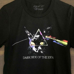 May include: Black t-shirt with a graphic of a cat's face in a triangle, with a rainbow prism effect behind it. The text "Dark Side of the Sofa" is printed below the graphic.