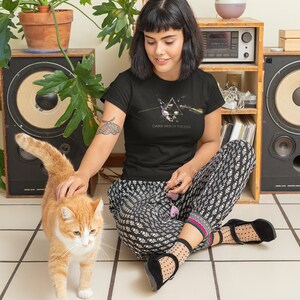 May include: A woman wearing a black t-shirt with a graphic of the Pink Floyd album cover "Dark Side of the Moon" and black and white patterned pants. She is sitting on a tiled floor and petting a ginger cat.