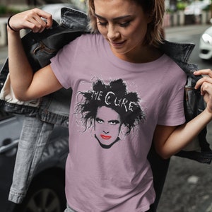 May include: A woman wearing a mauve t-shirt with a black and white graphic of Robert Smith from The Cure. The t-shirt has the band's name printed in white above the graphic.