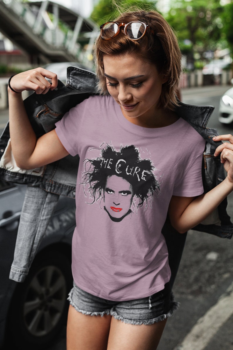May include: A woman wearing a mauve t-shirt with a black and white graphic of Robert Smith from The Cure. The t-shirt has the band's name printed in white above the graphic.