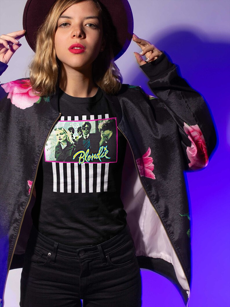 Op de afbeelding: Een vrouw draagt een zwarte bomberjack met roze bloemenprint, een zwart t-shirt met een band-afbeelding en de tekst "Blondie", en een zwarte jeans. Ze draagt een bordeauxrode fedora-hoed.