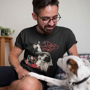 May include: A man wearing a black t-shirt with a red graphic of three dogs and the text "3 Dogs Strong". A white dog is sitting next to him.
