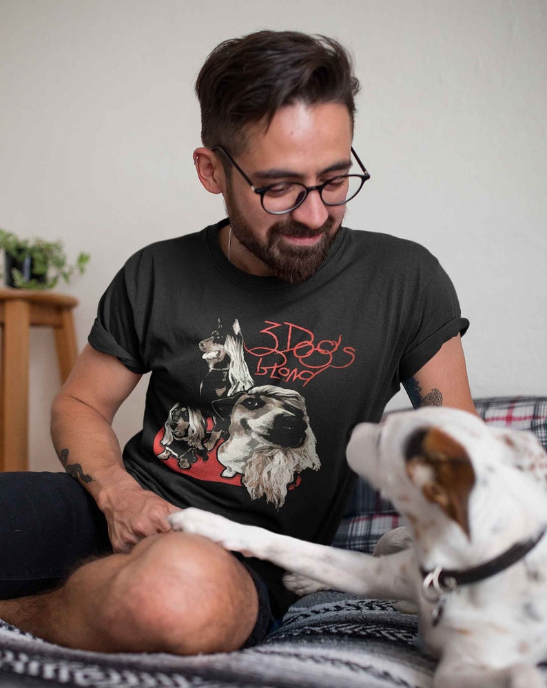 May include: A man wearing a black t-shirt with a red graphic of three dogs and the text "3 Dogs Strong". A white dog is sitting next to him.