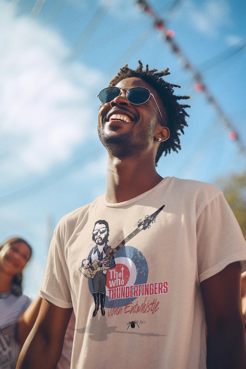 May include: A man wearing a white t-shirt with a cartoon illustration of John Entwistle from The Who playing a bass guitar. The shirt has the text "The Who Thunderfingers John Entwistle" printed on it.