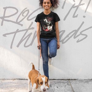 May include: A woman wearing a black t-shirt with a graphic of three dogs and the text "3 Dogs Night" is walking a brown and white beagle on a leash. The woman is standing in front of a white wall with the text "Rock It! Threads" written on it.